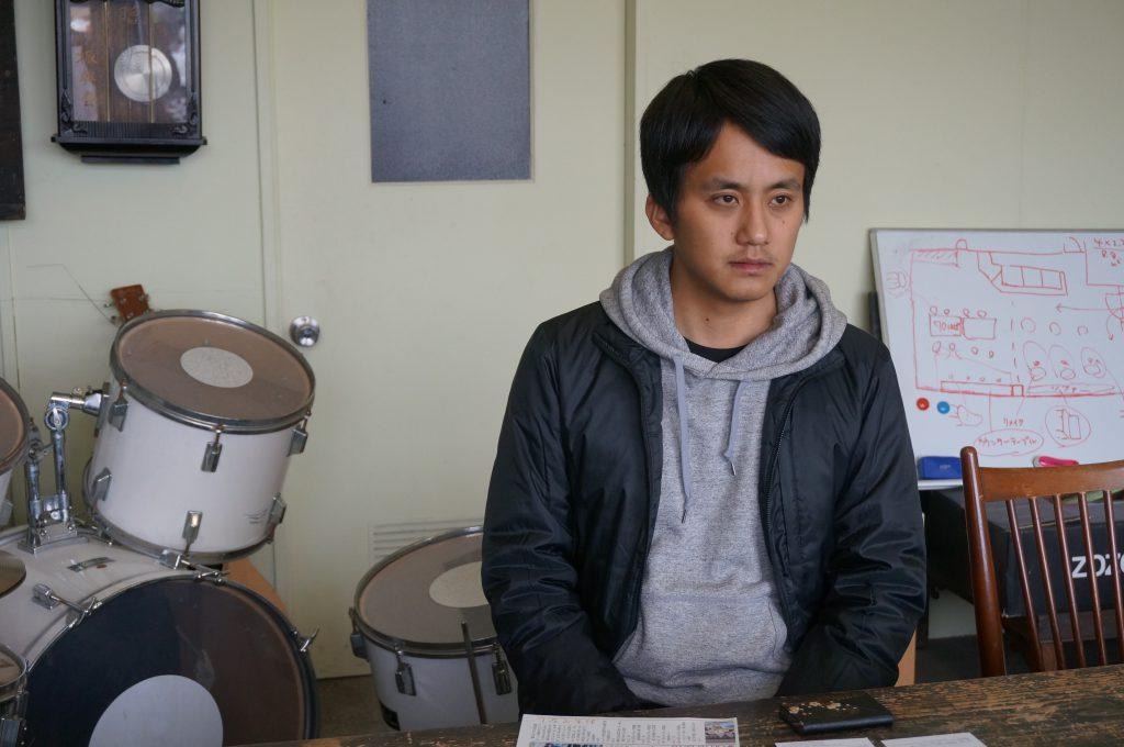 室内でインタビューに答える佐久田さんを写した写真、奥にはドラムセットと壁掛け時計がかけられたクリーム色の壁とガラス窓がついたクリーム色の開き戸と文字が書かれたホワイトボードが見える インタビューに答える佐久田さん