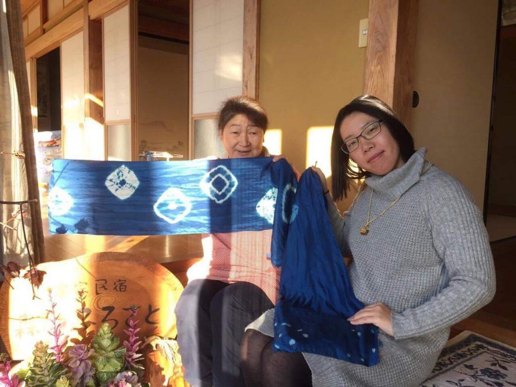 日本家屋の建物を背景に民宿ふるさとと書かれた木の看板の前で藍色の生地に白色の模様が入った織物を両手で広げた農家の女性と一緒に織物を持つ東山さんを写した写真 お世話になっている農家さんと