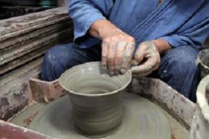 工房で、青い作務衣を着た職人が、両手で粘土の形を整えながら陶器を作っている写真