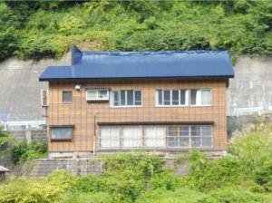 山の斜面と擁壁からなる山道を背景に、その山道沿いに建てられ茶色い木で横張りされた外壁と青い屋根で構成された大きな横長の家屋を、手前から撮影した写真