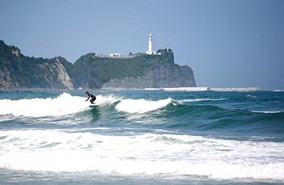 灯台の建つ崖を背に、サーフィンをする男性の写真