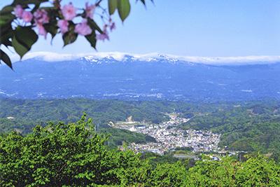 青空の下、手前には桜の花と新緑が美しく、また、遠くに連なる山々の頂には雪が積もり、麓には川俣町全体の様子が見える写真