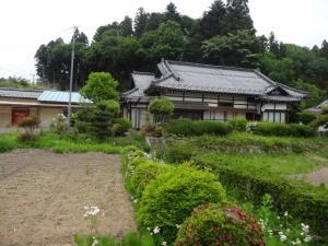瓦屋根の大きな日本家屋の写真。裏には林があり、玄関側には畑が2枚ある。