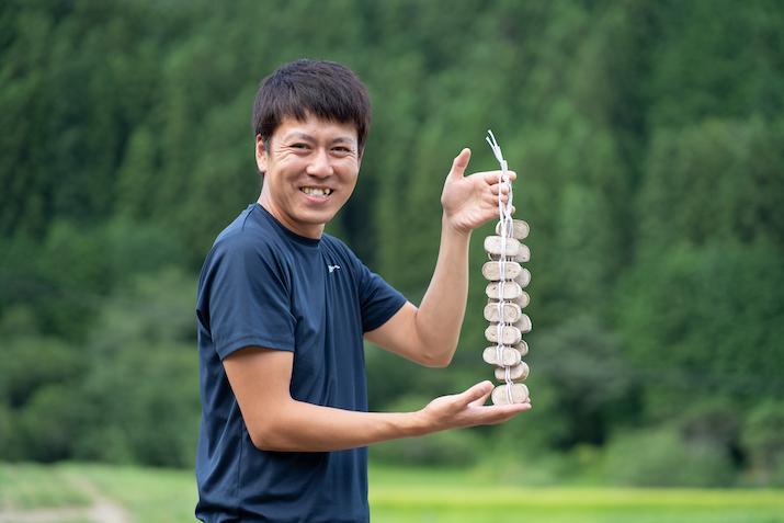 秋山健人さんが紐で吊るした凍み餅を笑顔で紹介している写真