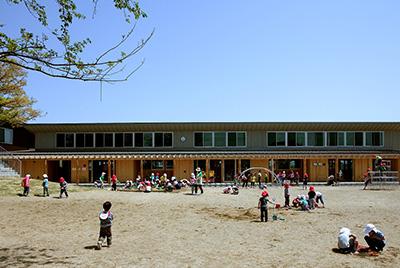 青空の下、施設のグラウンドで遊んでいる子供たちの写真。小さい子供たちが複数人いて、砂遊びをしている子供、遊具で遊ぶ子供などが写っている。