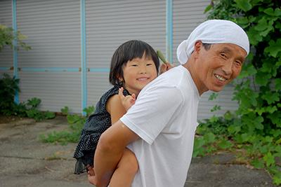 頭に白いタオルを巻いた中老の男性が、小さな女の子を背負い微笑みを浮かべている写真