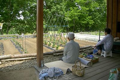 家の縁側に座り耕された庭の畑を眺める二人の男女の写真